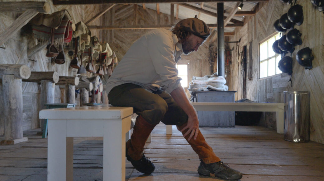 Screenshot Bearded man in a rustic workshop bends to adjust his boot, surrounded by riding gear and helmets.
