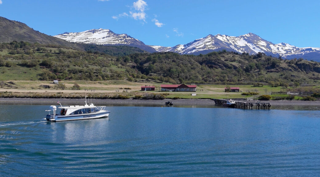 Screenshot A white tour boat crosses a blue lake beneath snow-capped mountains, with a small lakeside settlement and pier on shore.