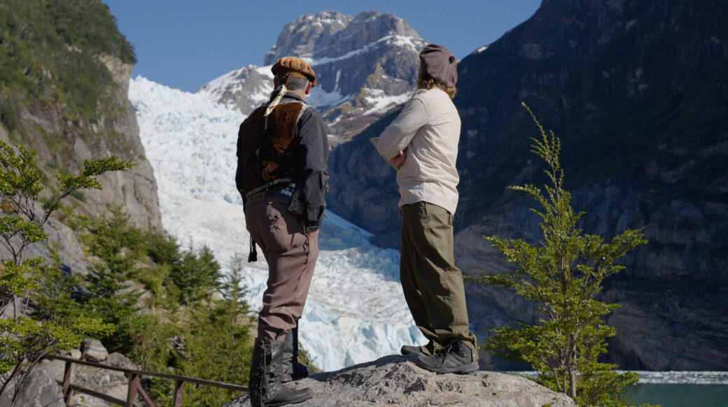 Screenshot Two hikers admire a massive glacier beneath snow-covered mountains in a rugged alpine valley.