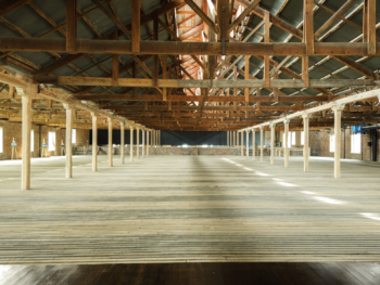 piso3 Vast empty warehouse interior with timber roof trusses, brick walls, wooden columns, and sunlight across the plank floor.