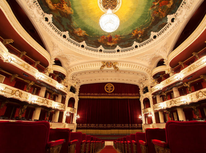 Ornate historic theater interior with red velvet seats, gilded balconies, a painted dome, and a glowing chandelier above the stage.