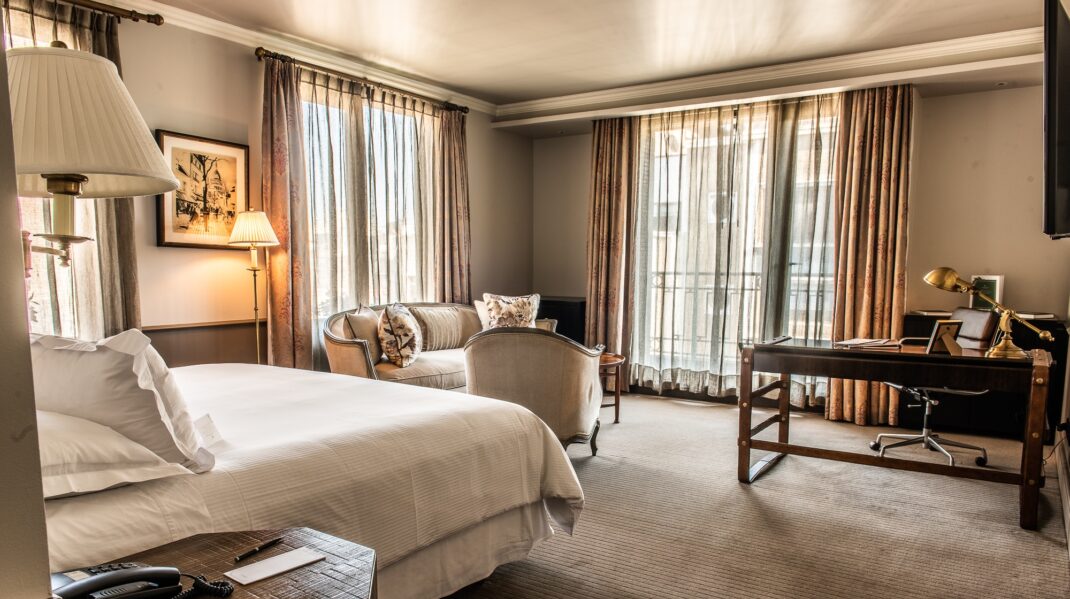 Elegant hotel suite with a king bed, sitting area, writing desk, and floor-to-ceiling curtained windows.