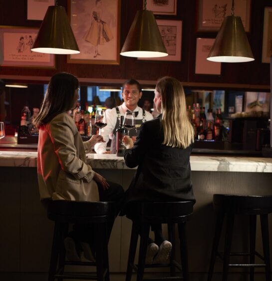Two women enjoy wine at a warmly lit bar while chatting with a smiling bartender.