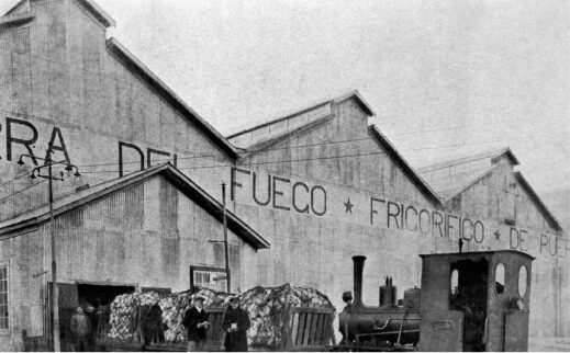 Black-and-white photograph of a steam locomotive and freight wagons outside a large Spanish railway warehouse labeled “Depósito de Fuego y Frigorífico.”