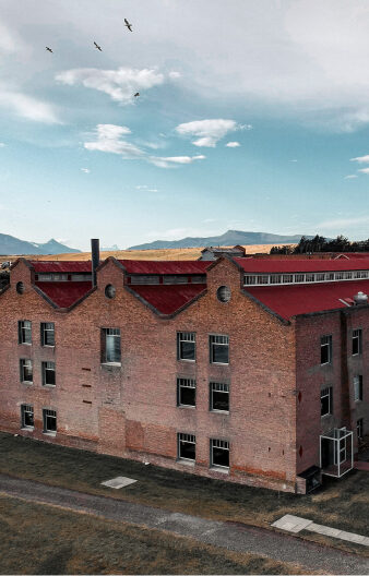 Large red-brick industrial building with a red roof beneath a cloudy blue sky, birds flying overhead and mountains in the distance.