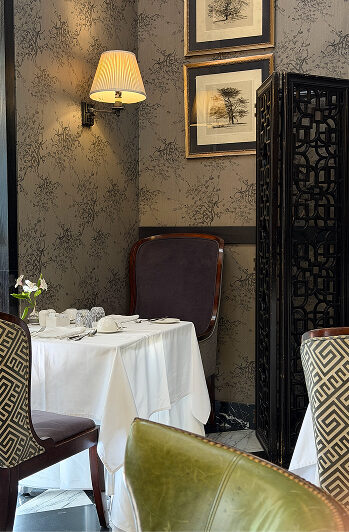 Elegant restaurant dining nook with patterned wallpaper, framed artwork, wall sconce, white tablecloth, and upholstered chairs.