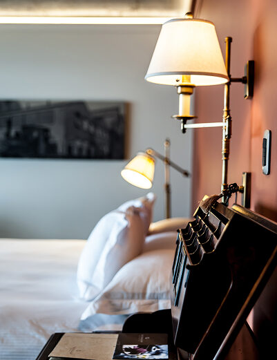 Elegant hotel bedroom with a bedside desk, open book, white pillows, and warm wall-mounted lamps.