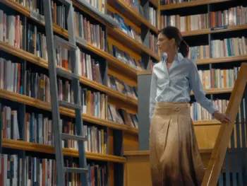 the-singular-libreria-wordpress Woman browsing books in a library, standing beside a wooden rolling ladder and tall bookshelves.